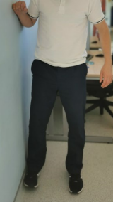 A man standing against a wall lifting one leg sideways, keeping his foot against the wall, and holding onto the wall with his arm for balance. The other foot is flat on the floor.