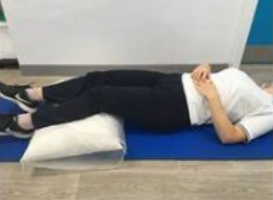 A person lying on their back with their hands placed on their stomach and their legs raised slightly by a cushion placed under their calves and knees.