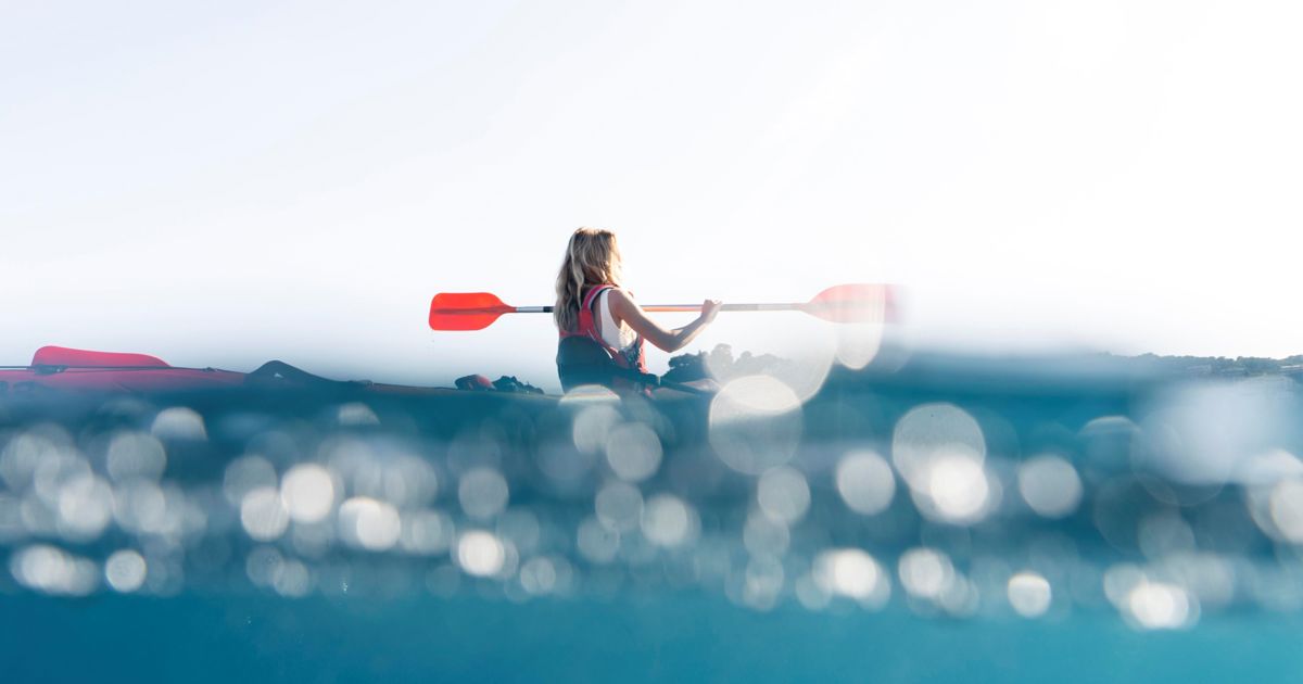 A young blonde woman in a life jacket is sitting in a canoe with the sea rippling around her. she holds orange ores and is paddling with them. 