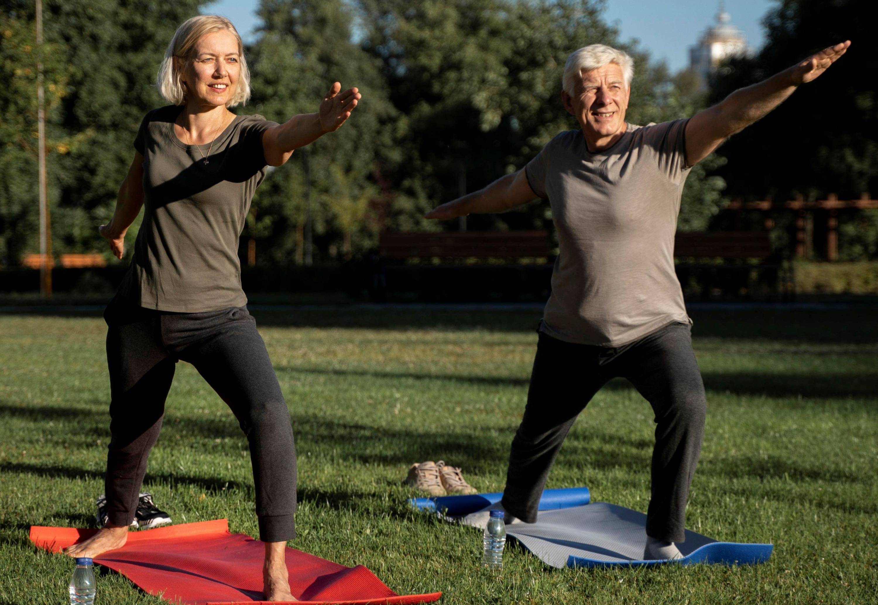 A senior couple on yoga mats outside in a park in a bent knee standing pose with arms outstretched.