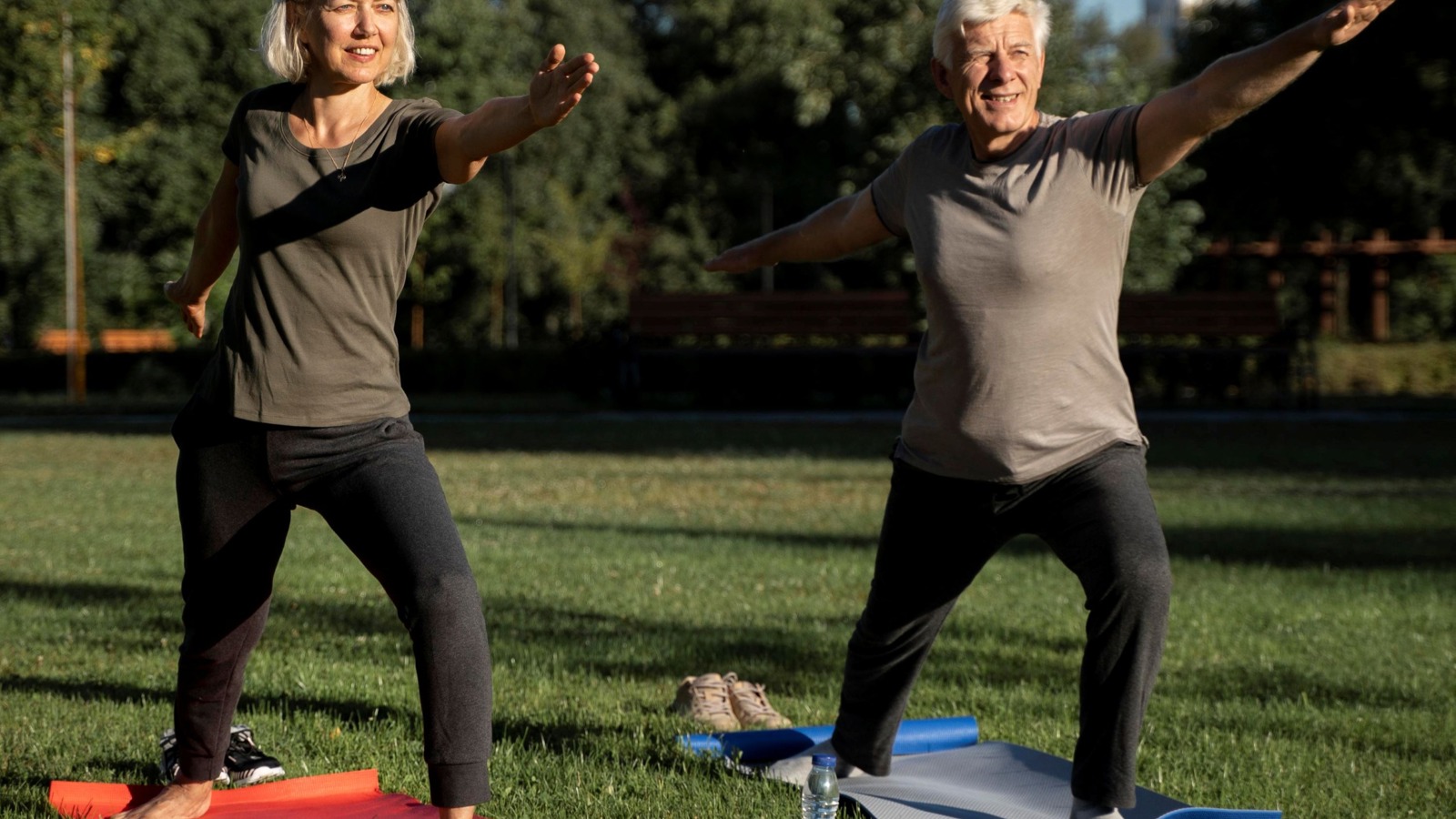 A senior couple on yoga mats outside in a park in a bent knee standing pose with arms outstretched.