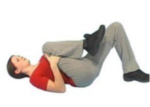 a yung woman lays on the floor on her back with one knee bent and the other knee raised up to her chest. Her hands hold the pose to stretch out her back. 