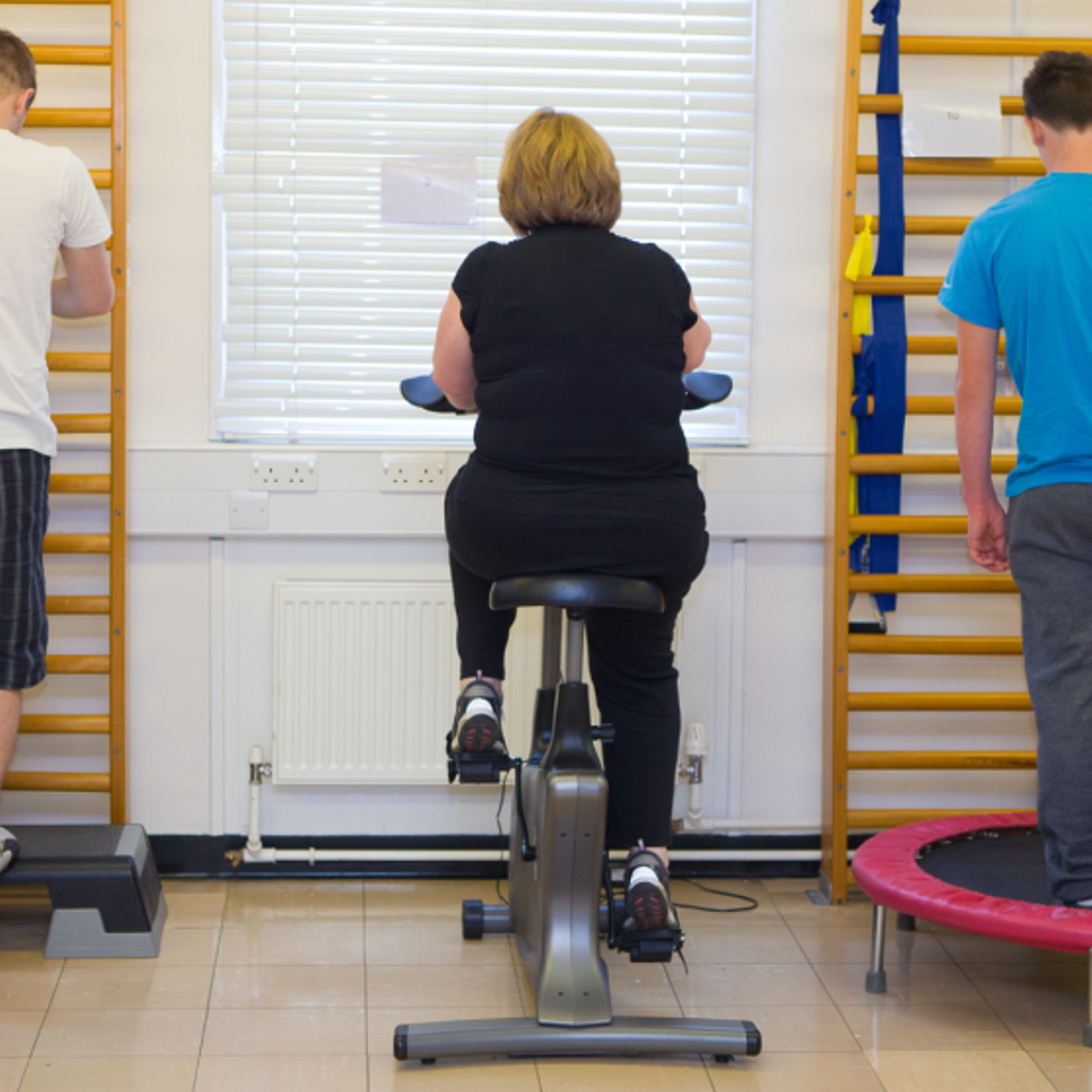 A photograph of a gym based rehabilitation session with 3 patients with their backs to the camera. one man step sup onto a step, next to an  elderly woman on an exercise bicycle and finally a young man jumping on a trampette, all in a row.. 