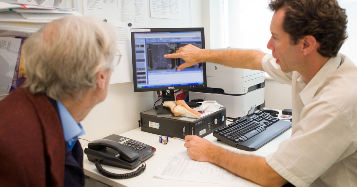 Clinician Sitting At Desk Next To A Patient And Pointing At His Computer Screen
