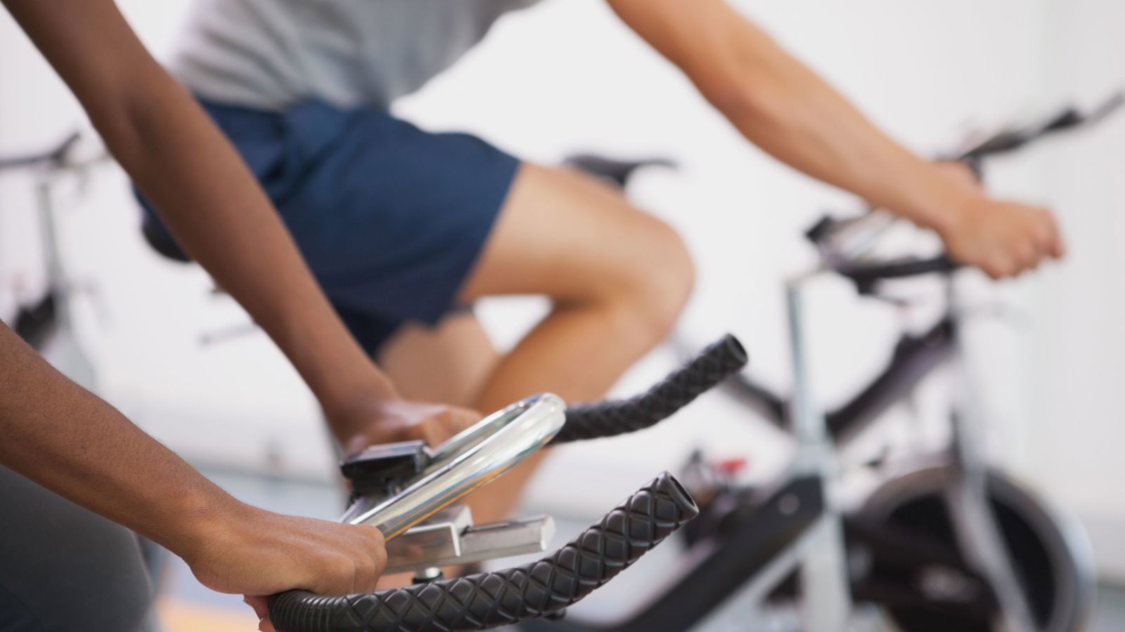 cropped image of two people, one black and one while in shorts and t-shirts on an exercise bike pedalling. Close crop on their hands on handle bars and their legs moving. 